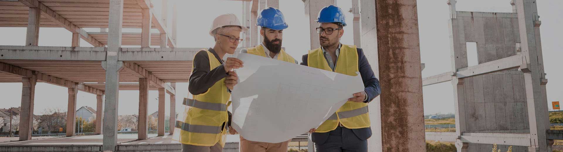 Two business woman and one man wearing hard hats in front of a construction site.